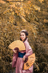 Obraz premium Japanese geisha in a traditional kimano with a fan and armed with a katana sword in a beautiful garden. A girl from medieval Asia. Reconstruction of cultural heritage. Culture in Japan.