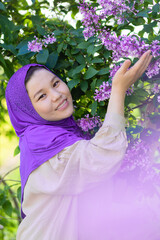 beautiful young asian islamic woman in hijab, headscarf sniffing lilac. happy muslim Kazakh girl with spring summer flowers. vertical