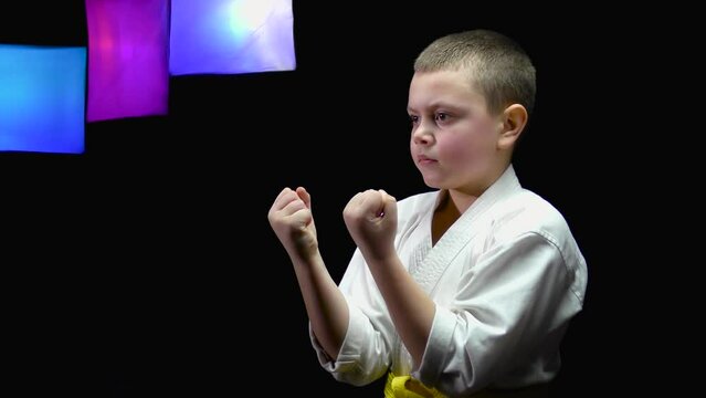 A boy athlete with a yellow belt trains punches on a black background with color-changing spotlights