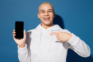 Young dyed blond man of African American ethnicity in white hoody hold use point finger on mobile cell phone with blank screen workspace area isolated on plain dark royal navy blue background studio