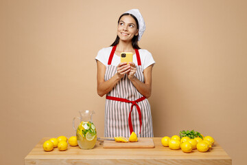 Obraz premium Young fun housewife housekeeper chef latin woman wear apron toque hat work at table kitchenware cutting lemons make lemonade use mobile cell phone look aside isolated on plain pastel beige background.