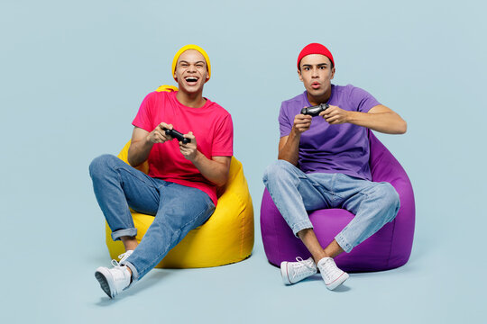 Full body young couple two friends men in casual clothes together sit in bag chair hold in hand play pc game with joystick console isolated on pastel plain light blue cyan background studio portrait