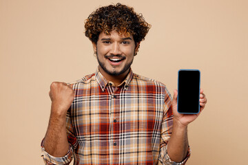 Obraz na płótnie Canvas Young Indian man wear brown shirt casual clothes hold in hand use mobile cell phone with blank screen workspace area do winner gesture isolated on plain pastel light beige background studio portrait.