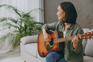 Side view young smiling happy calm woman of Asian ethnicity wearing casual clothes play guitar sits on grey sofa couch stay at home hotel flat rest relax spend free spare time in living room indoor