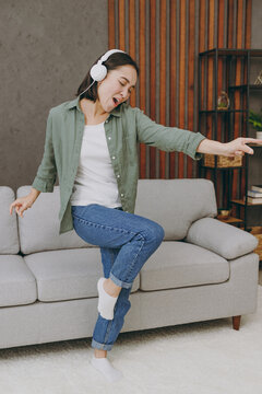 Full body young woman of Asian ethnicity wears casual clothes headphones listen dancing stand near grey sofa dance couch stay at home hotel flat rest relax spend free spare time in living room indoor.