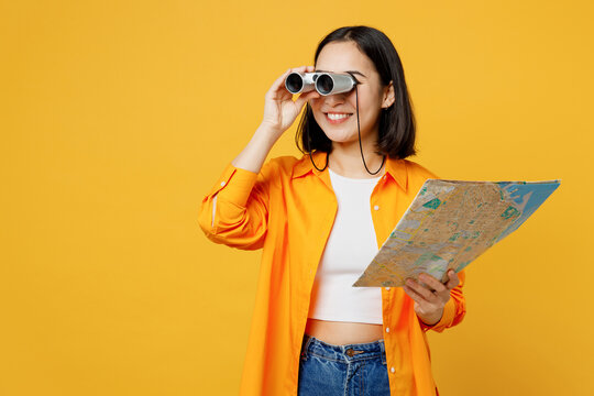 Young happy woman wear summer casual clothes hold in hand map use binoculars isolated on plain yellow background. Tourist travel abroad in free spare time rest getaway Air flight trip journey concept.