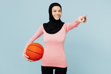 Young arabian muslim fitness trainer sporty woman wear pink abaya hijab spend time in home gym hold basketball ball point aside isolated on plain blue background studio Workout sport fit abs concept.