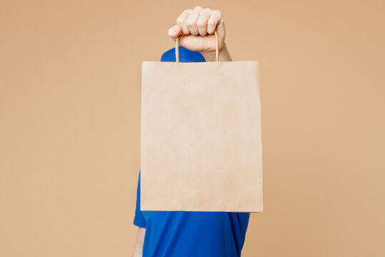 Delivery guy employee man wear blue cap t-shirt uniform workwear work as dealer courier cover face with hold brown clear blank craft paper takeaway bag mockup isolated on plain light beige background