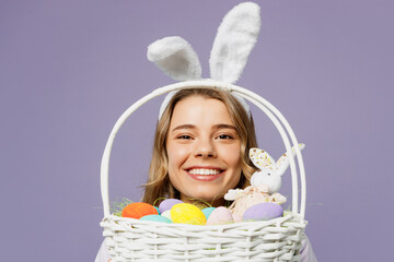 Close up young smiling fun woman wearing bunny rabbit ears hold wicker basket colorful eggs look aside on area isolated on plain pastel light purple background studio portrait. Happy Easter concept.