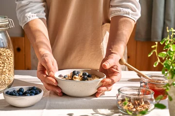 Foto op Canvas Woman preparing healthy dieting vegan nutritious breakfast. Female hands holding bowl of oatmeal porridge with blueberries, walnuts and honey. © Caterina Trimarchi