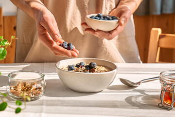 Woman preparing healthy dieting vegan nutritious breakfast. Female hand putting blueberries in the bowl with oatmeal porridge with walnuts and honey.