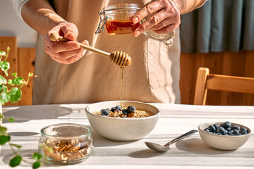 Woman preparing healthy dieting vegan nutritious breakfast. Female hand pouring honey in the bowl with oatmeal porridge with walnuts and blueberries. - obrazy, fototapety, plakaty