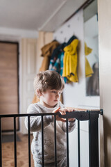 Front view of a little boy standing on the other side of the child safety gate. A little boy is holding on to the top of the security gate and looking at the camera from above.