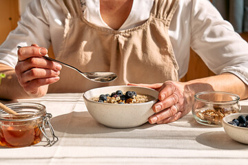 Woman eating healthy dieting vegan nutritious breakfast. Female hand holding spoon over the bowl with oatmeal porridge with walnuts, blueberries and honey. - obrazy, fototapety, plakaty