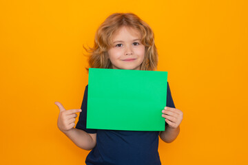 Fototapeta na wymiar Child pointing on empty green sheet of paper, isolated on yellow background. Portrait of a kid holding a blank placard, poster. Mockup, copyspace.