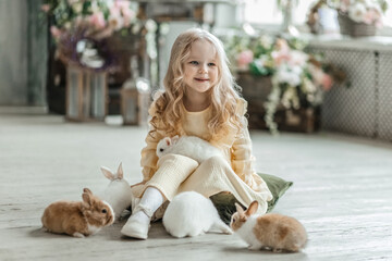 A little smiling girl in a yellow dress is playing on the floor in a room with rabbits. A baby and a rabbit. The living room is decorated for Easter.