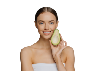 Horizontal shot of happy brunette woman holds half of avocado near face uses anti aging organic recipes stands refreshed indoor wrapped in towel isolated over blue background. Healthy diet concept
