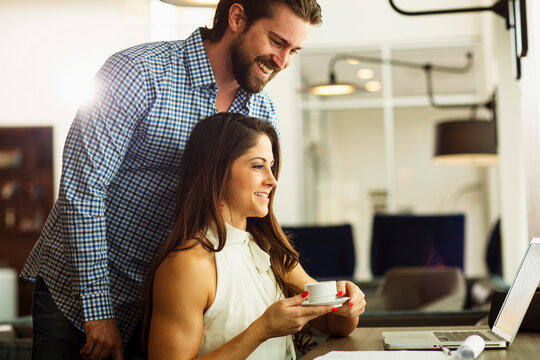 A Young Beautiful Lady Sits On A Chair With A Cup Of Tea On Her Both Hands, Looks At Her Laptop That Is On A Table Before Her.  Her Male Partner Stands Behind Her Too Looks At Her 