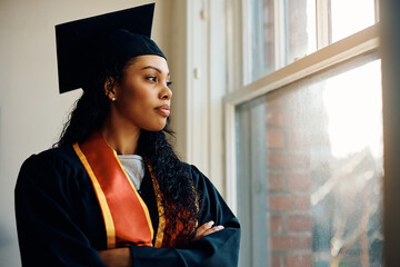 Pensive African American woman in graduation gown looks through window.