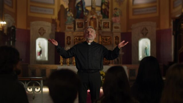 A gray-haired Catholic priest conducts a Sunday service. The priest throws up his hands and looks up while praying.