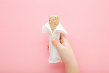 Baby girl hand holding halva bar on light pink table background. Pastel color. Opened white pack....