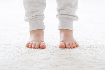 Little child legs in white warm trousers standing on light beige carpet at home room. Barefoot...