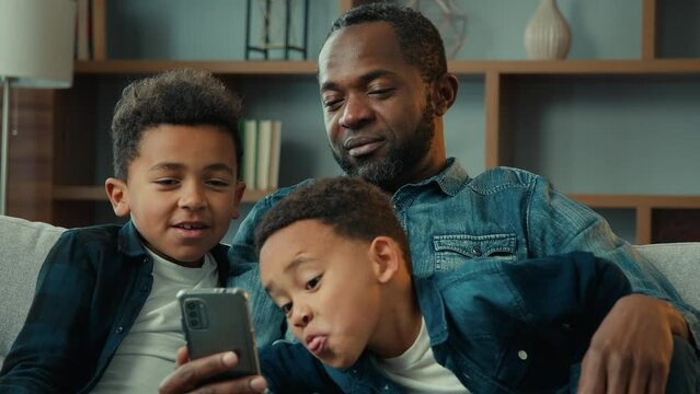 African American family father man and little kids sons boys children sit on sofa look at mobile screen having fun using phone try funny app photo mask taking selfie grimace looking cellphone at home