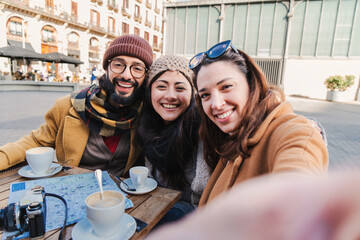 Group of young tourist people smiling and having fun taking a selfie portrait looking at camera. Front view of multiracial friends doing a photo in a coffee shop or restaurant terrace. Lifestyle