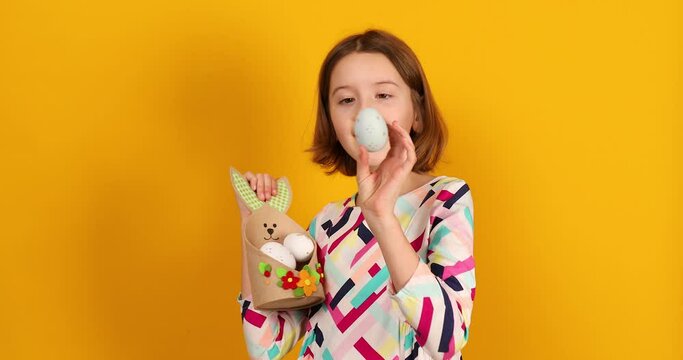 Happy playful teenager girl with painted Easter eggs on basket like a bunny, on a bright yellow studio background, celebrating Easter in style.
