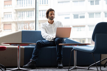 portrait d'un jeune homme assis au bureau dans un open space devant son ordinateur portable....