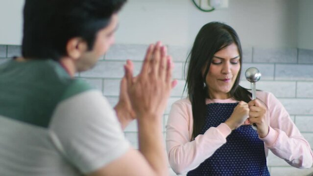 Young happy Indian Asian wife wearing a Chef Apron singing using a ladle and smiling husband appreciates her in a modern kitchen house setup. Bonding, relationship, family, gender equality concept