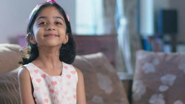 A playful young happy Indian asian little girl kid or female child smiling and looking at the camera sitting on a couch indoors in an interior house setup.