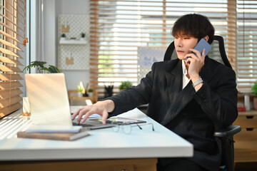 Young asian male business executive looking at laptop screen, discussing project on mobile phone