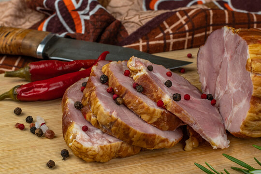 Sliced slices of juicy pork ham on a wooden cutting board, next to it are a carving knife, multi-colored allspice peas, red hot peppers, parsley, dill and basil. The concept of delicious meat products