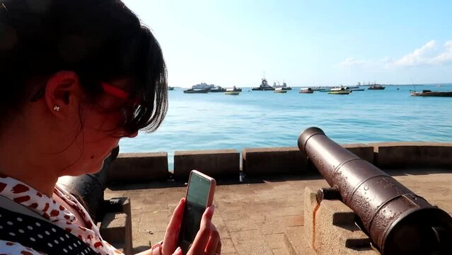 Woman filming with the phone some old canyons in front of some boats navigating in the Ocean. Stone Town, Zanzibar