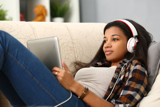 Happy african american young woman lying on sofa with tablet computer and headphones listening to music at home