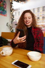 Portrait of a red-haired girl holding a cup of coffee and taking a photo with a smartphone. She is looking at the phone camera and smiling