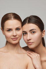 Closeup portrait of two adorable young girls with well-kept skin posing over grey background. Models with bare shoulders. Concept of beauty, spa, skin care and health