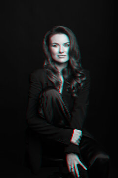 Woman portrait with black suit and big wavy hair in red and blue color split effect style. Model sitting on chair and hugging her one leg. Woman looking at camera. Futuristic looking style
