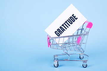 White blank card with GRATITUDE text on shopping trolley on light blue background.