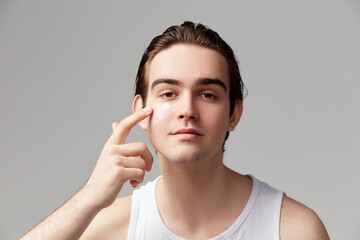Portrait of young handsome boy applying moisturizing face cream, mask, posing against grey studio...