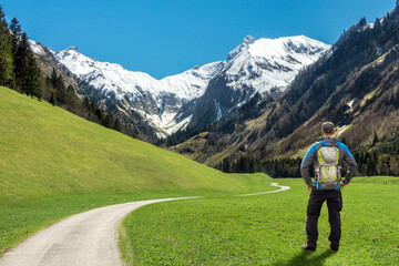 Hiker Man with Backpack standing on green grass meadow and hiking trail enjoying snowcapped mountains in springtime. Trettachtal, Allgaeu, Bavaria, Germany. - 577011766