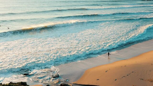 Surfer with surfboard staying on the beach and watching waves. Ocean and sandy coast in warm sunset light, view from above.