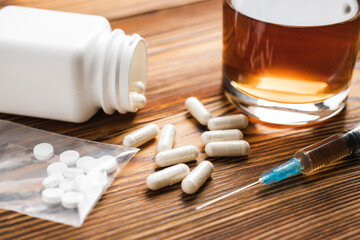 White pills, syringe with a drugs dose, narcotics, glass with alcohol drink on a wooden table, close-up view. Concept of addiction and bad habits