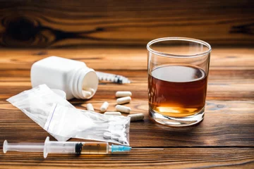 Glass with alcohol drink, whisky or brandy, white pills, syringe with a drugs dose, narcotic in transparent bag on a wooden table. Concept of addiction and bad habits © O.Farion