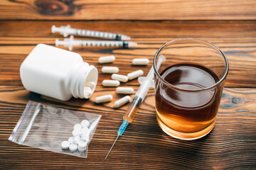 Glass with alcohol drink, whisky or brandy, white pills, syringe with a drugs dose, narcotic in transparent bag on a wooden table. Concept of addiction and bad habits