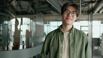 Smiling brunet Asian man wearing eyeglasses waving hand at the camera in a co-working room