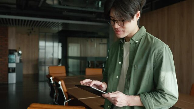 Smiling Asian man in eyeglasses working on tablet and looking at window in a co-working room