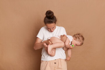 Photo of mom looking in stinky diaper of her little daughter, holding infant kid and does a grimace for disgust, standing isolated over brown background. - obrazy, fototapety, plakaty