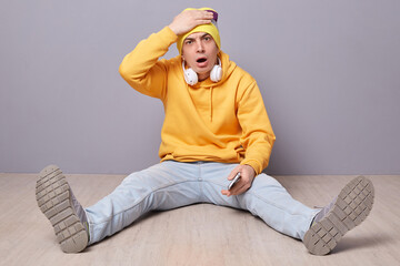 Portrait of shocked disappointed man wearing beanie hat, yellow hoodie and jeans posing against gray wall, sitting on floor, making facepalm gesture, forgot something important.